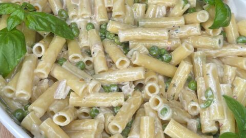 A white dish filled with creamy rigatoni pasta mixed with green peas, garnished with fresh basil leaves, sitting on a wooden surface.