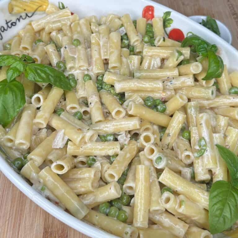 A white dish filled with creamy rigatoni pasta mixed with green peas, garnished with fresh basil leaves, sitting on a wooden surface.