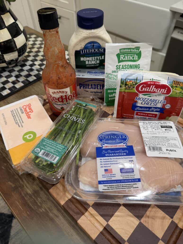 A wooden board holds chicken breasts, cheddar cheese, chives, mozzarella cheese, ranch dressing, hot sauce, dry ranch seasoning, and cream cheese in a kitchen setting.