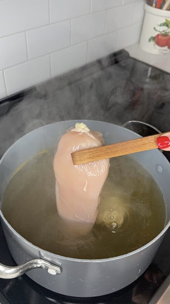 A hand with red nail polish holds a raw chicken breast with a wooden spoon over a steaming pot of hot water on a stove.