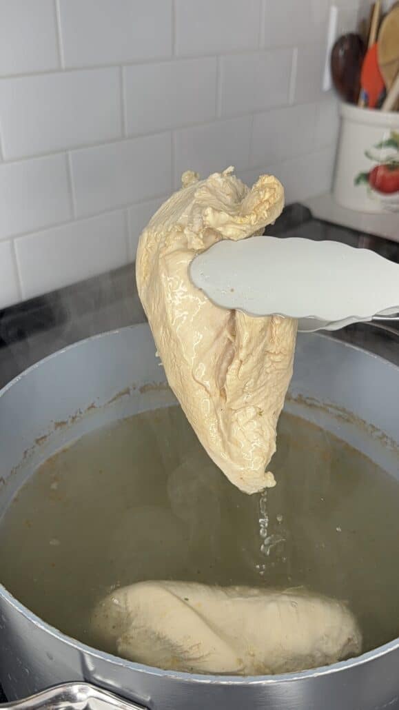 Tongs hold a piece of cooked chicken breast over a pot of simmering water on a stove. Another piece of chicken remains in the pot. Kitchen utensils are visible in the background.