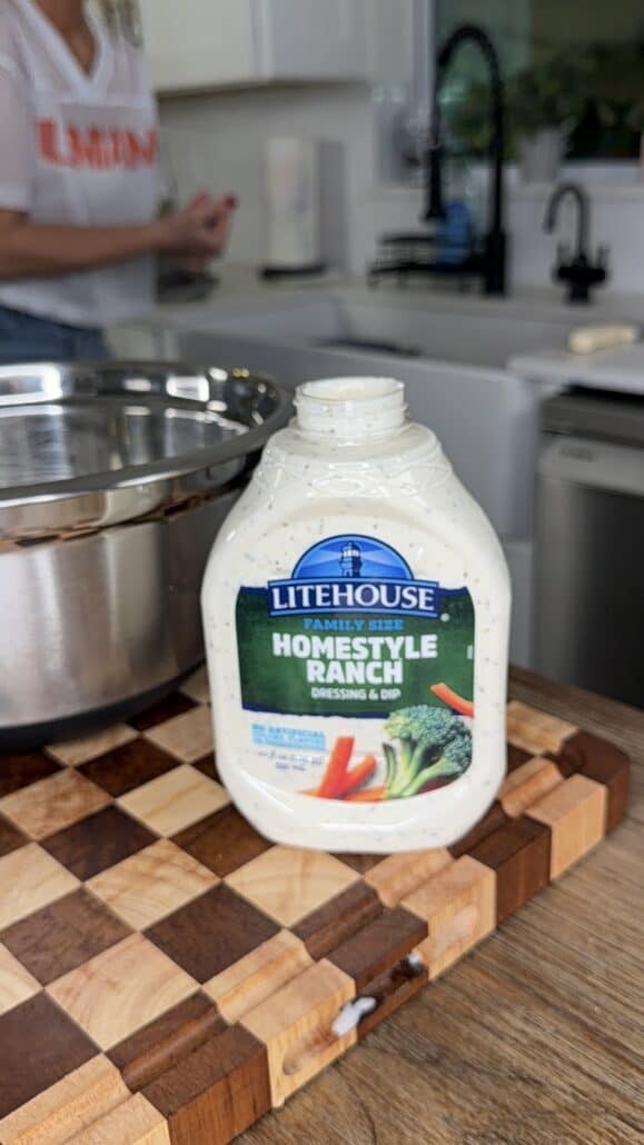 A bottle of Litehouse Homestyle Ranch dressing sits on a wooden cutting board next to a metal bowl, with a person blurred in the background near a white kitchen sink.
