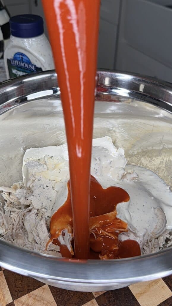 Buffalo sauce is being poured into a metal bowl containing shredded chicken, cream cheese, and ranch dressing. A jar of ranch is visible in the background on a kitchen counter.