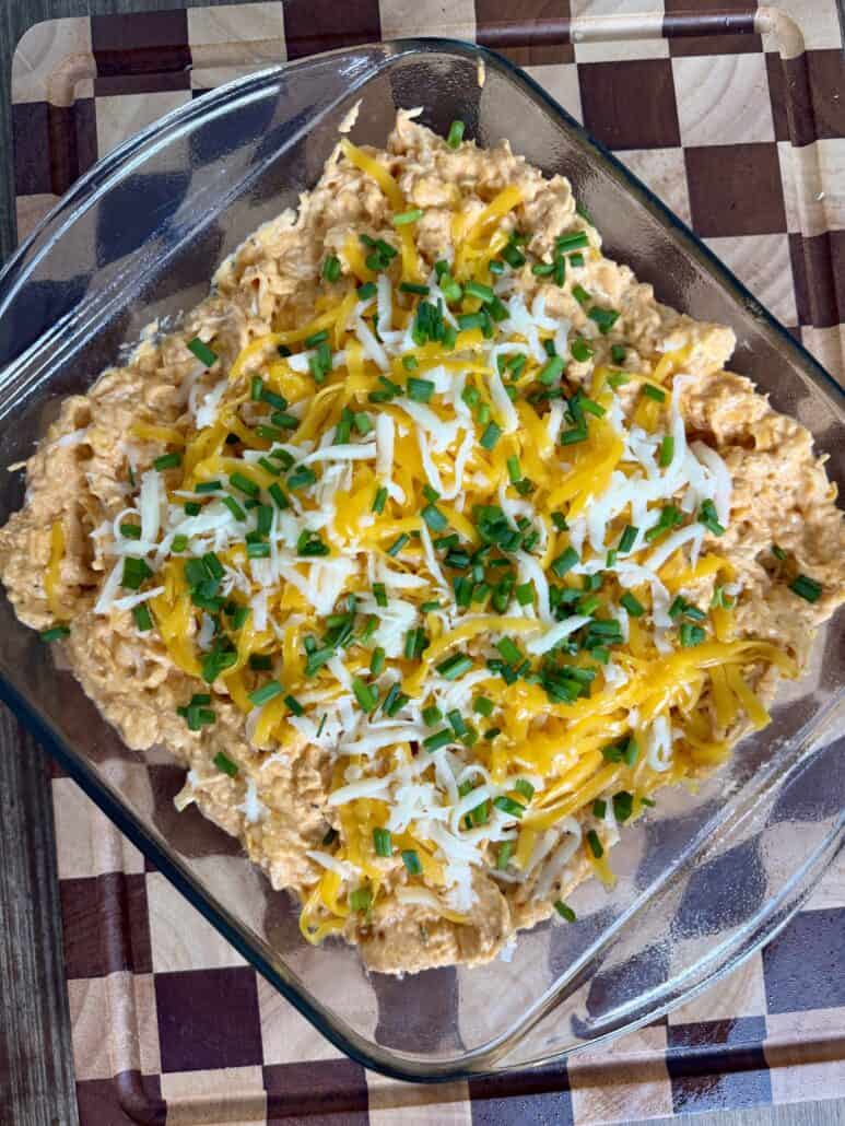 A glass dish of cheesy buffalo chicken dip topped with shredded cheese and chopped chives, sitting on a checkered wooden cutting board.