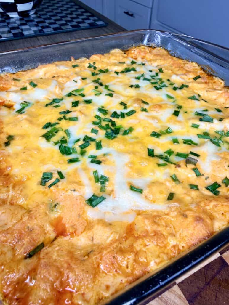 A baked casserole topped with melted cheese and chopped green onions sits in a glass dish on a kitchen counter.