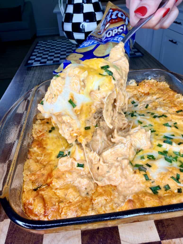A hand scoops cheesy buffalo chicken dip from a glass baking dish, with a bag of chips and a checkerboard pattern in the background.