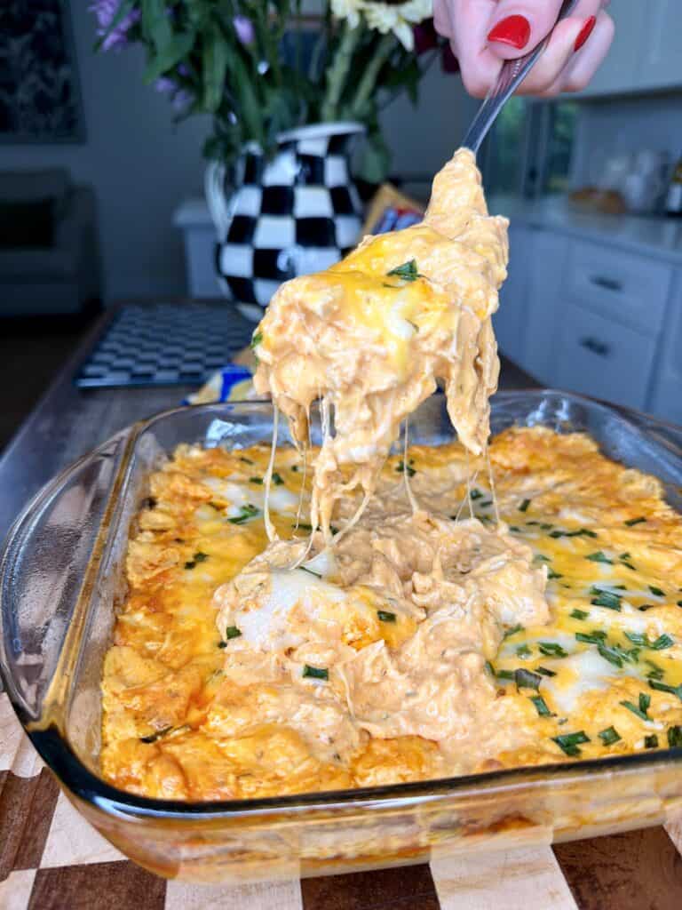 A hand with red nail polish lifts a cheesy, creamy buffalo chicken dip from a glass baking dish, with melted cheese stretching up. The dish sits on a wooden surface in a bright kitchen.