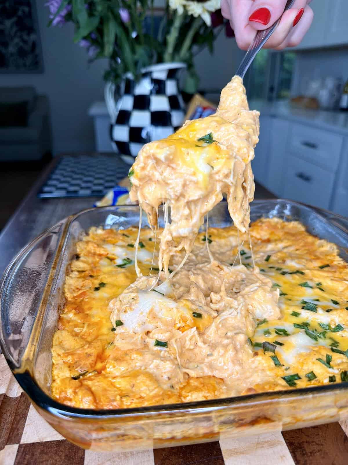 A hand with red nail polish lifts a cheesy, creamy buffalo chicken dip from a glass baking dish, with melted cheese stretching up. The dish sits on a wooden surface in a bright kitchen.
