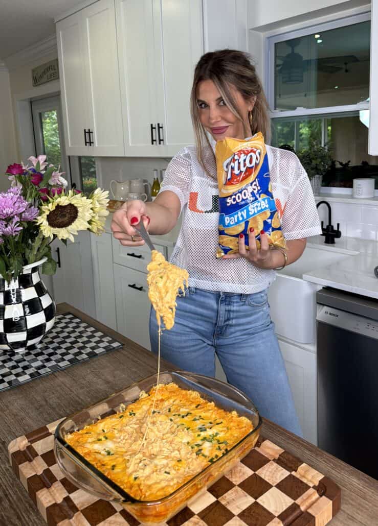 A woman in a kitchen smiles while holding a bag of Fritos and lifting cheesy dip from a glass dish with a chip. Flowers and a checkered vase are on the counter beside her.