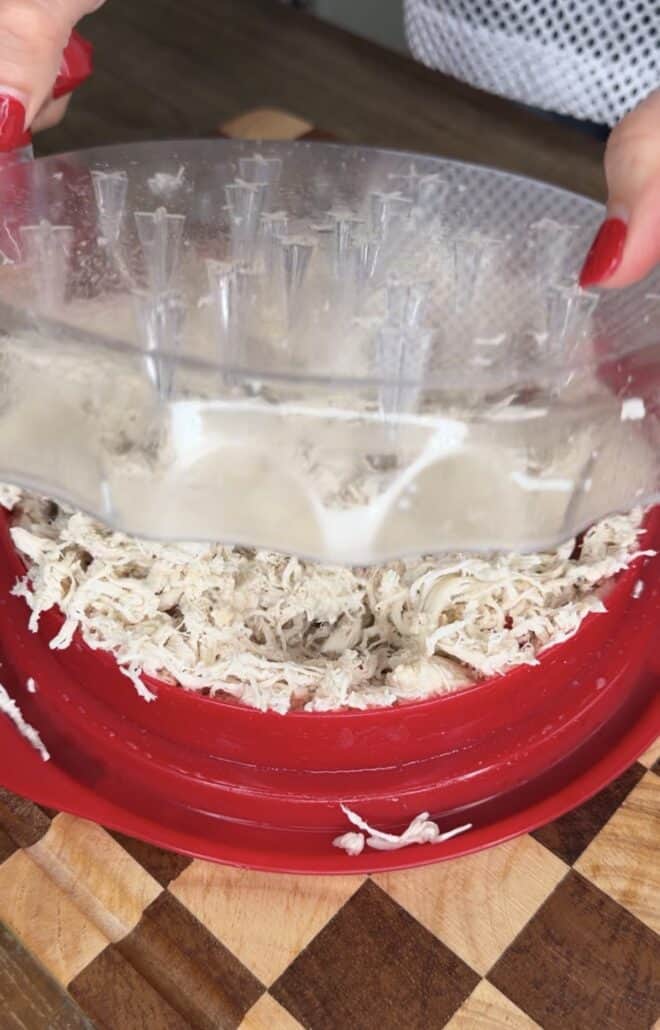A person uses a clear plastic tool to shred cooked chicken in a red bowl on a wooden surface.