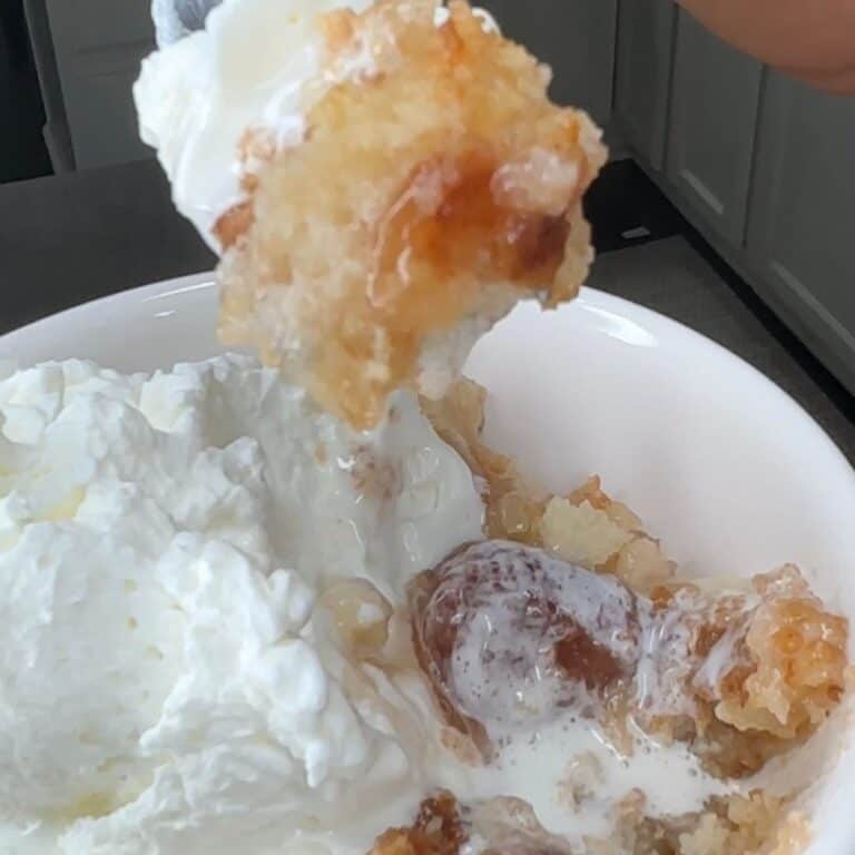 A close-up of a spoonful of warm apple cobbler topped with whipped cream being lifted from a white bowl filled with more cobbler and cream.