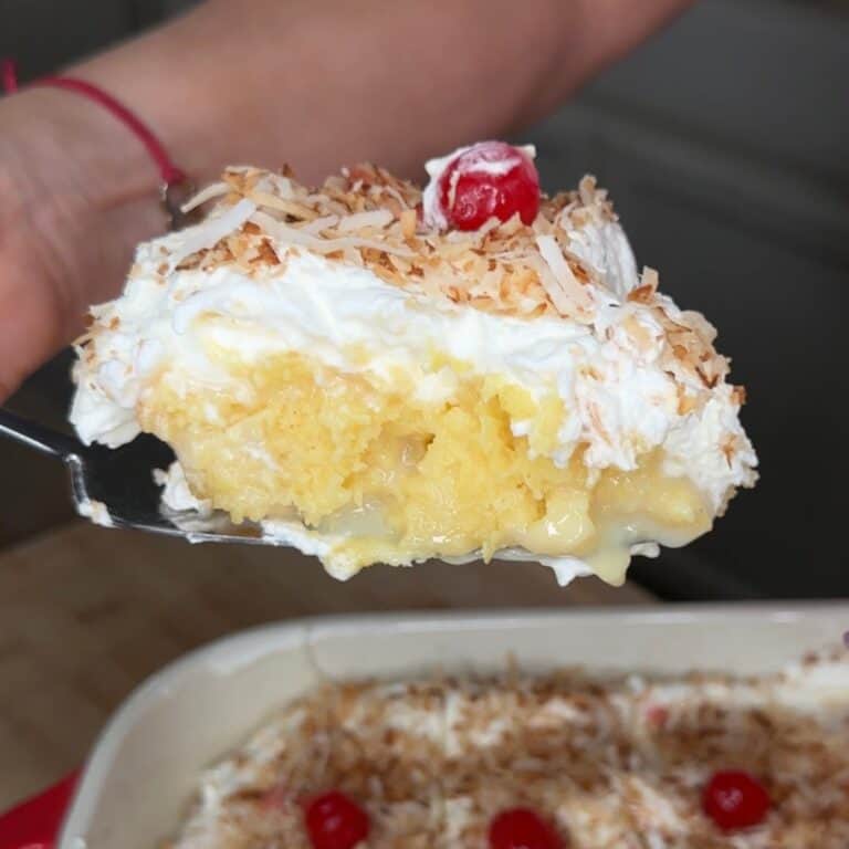 A close-up of a slice of creamy yellow cake topped with whipped cream, toasted coconut flakes, and a cherry, being lifted from a baking dish with more cake, coconut, and cherries.