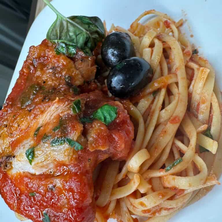 A plate of pasta with tomato sauce, garnished with basil leaves, black olives, and a piece of baked chicken topped with tomato sauce and herbs.
