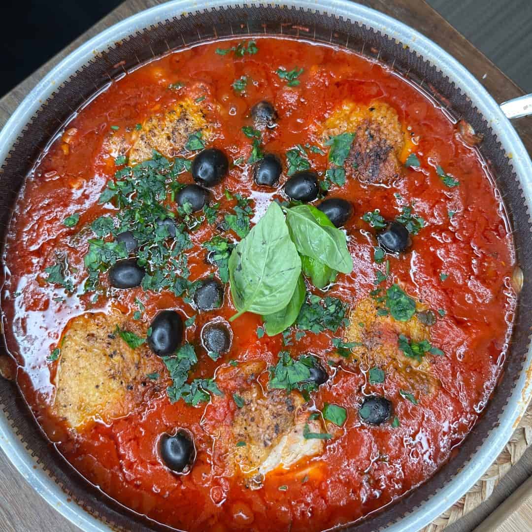 A pan filled with tomato sauce, baked chicken pieces, black olives, chopped parsley, and fresh basil leaves, all garnished on top.