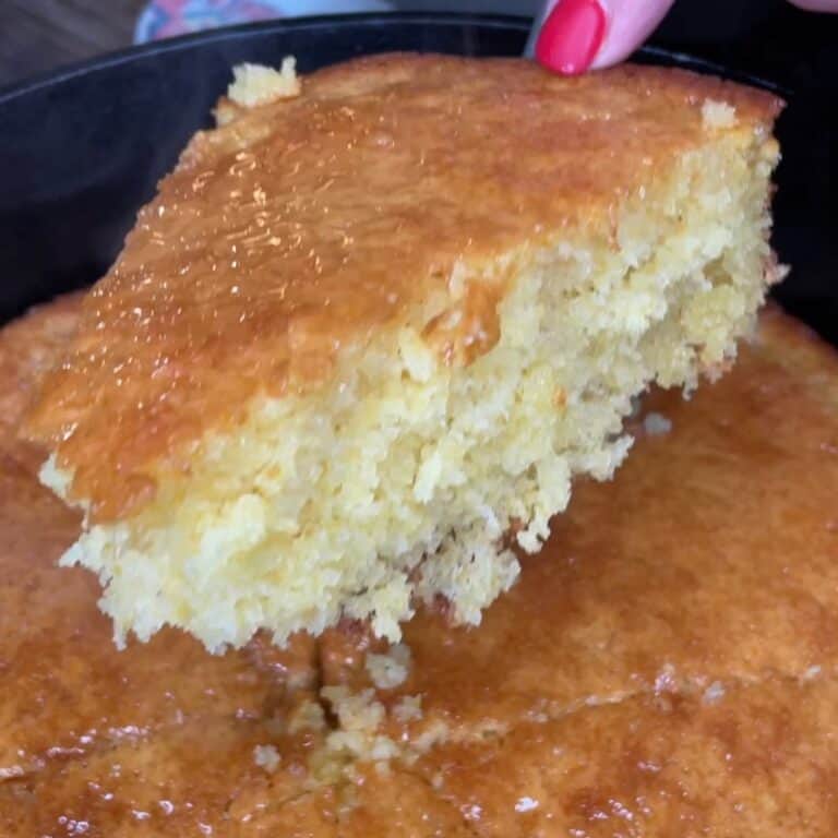A close-up of a hand with red nail polish holding a moist, golden slice of cornbread above a pan filled with the rest of the cornbread. The surface looks shiny and slightly crumbly.