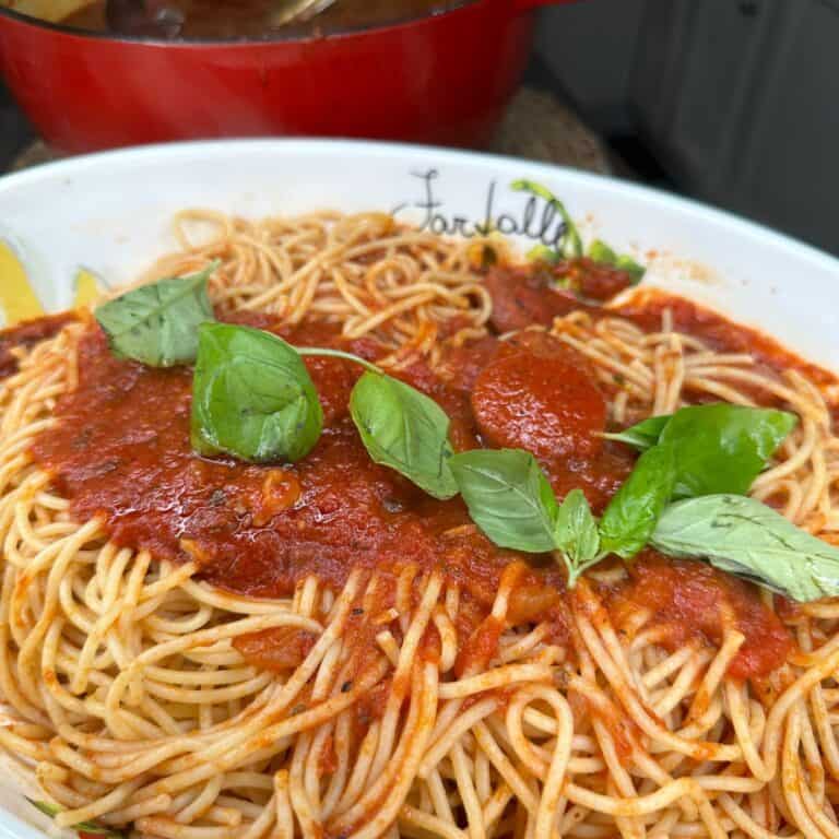 A plate of spaghetti topped with tomato sauce, pepperoni slices, and fresh basil leaves, served in a white dish with a red pot visible in the background.