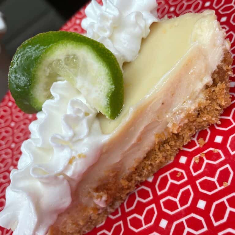A slice of key lime pie with a graham cracker crust, topped with whipped cream and a lime slice, served on a red plate with a white geometric pattern.