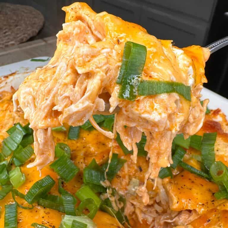 A close-up of a cheesy buffalo chicken casserole topped with sliced green onions, with a portion being lifted out on a spatula, showing melted cheese and shredded chicken.
