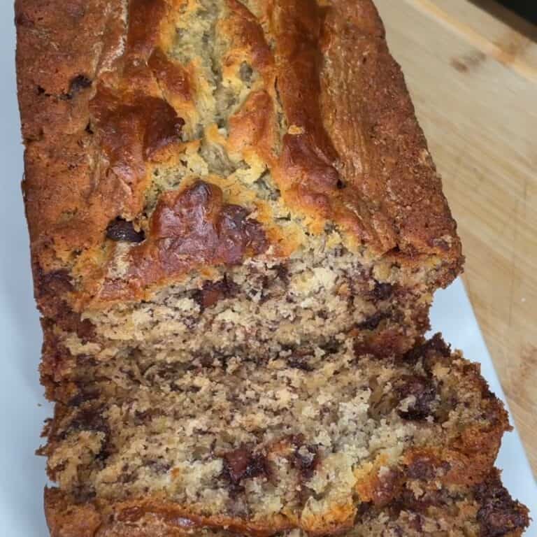 Close-up of a freshly baked loaf of banana bread with chocolate chips, partially sliced to reveal its moist, dense texture and visible chocolate pieces inside. The bread rests on a wooden surface.