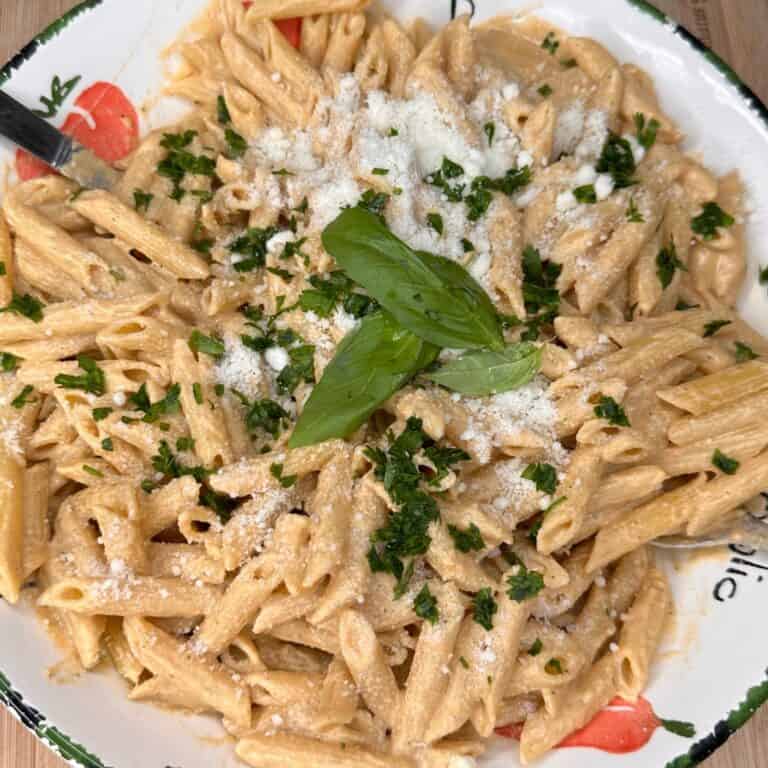 A plate of penne pasta in a creamy sauce, topped with grated cheese, chopped parsley, and a sprig of fresh basil in the center. A fork rests on the side of the dish.