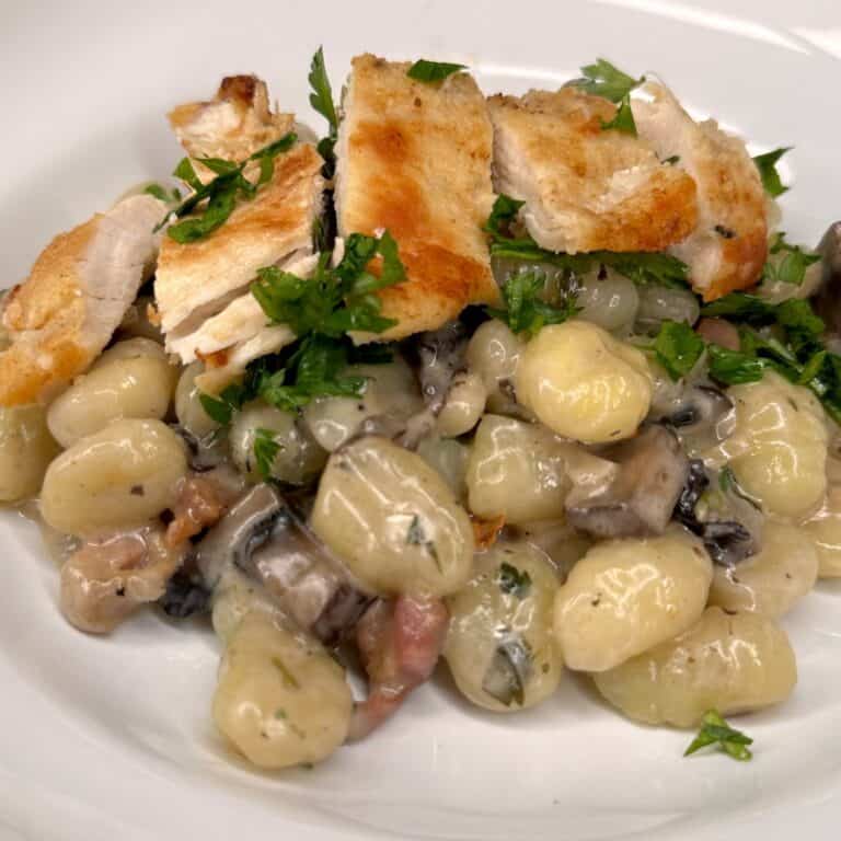 A plate of creamy gnocchi with mushrooms and bacon, topped with sliced grilled chicken and garnished with fresh chopped parsley.