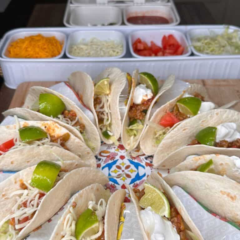 A circular arrangement of soft tacos filled with ground meat, shredded cheese, lettuce, tomato, sour cream, and lime wedges. In the background are containers of taco toppings, including cheese, tomatoes, and sauces.