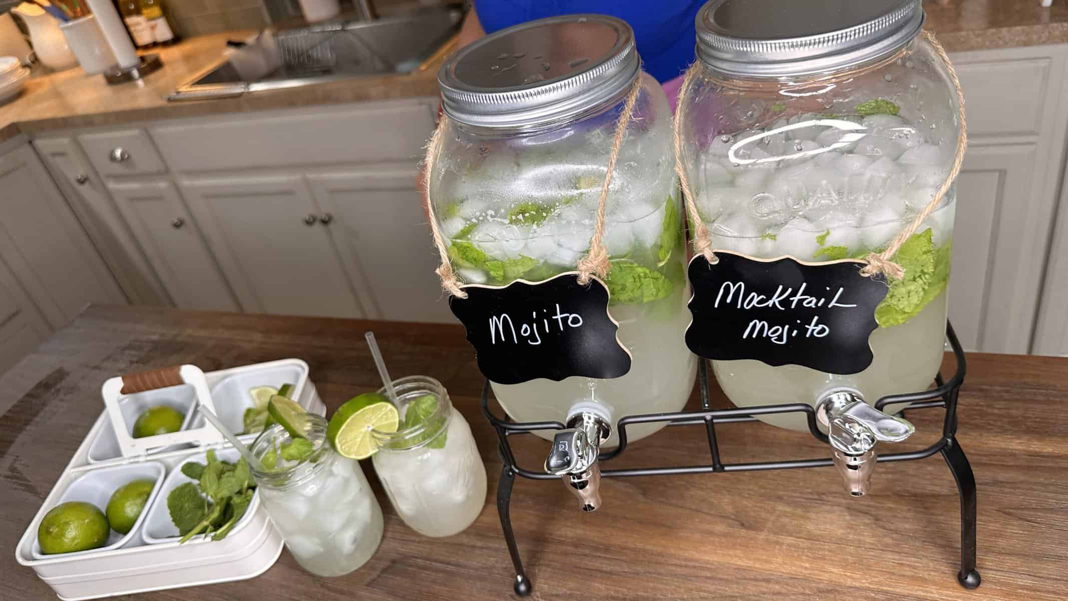 Two large glass dispensers labeled โMojitoโ and โMocktail Mojitoโ sit on a counter, filled with ice, lime, and mint. In front, two mason jars with drinks, lime slices, and a tray with limes and mint are visible.