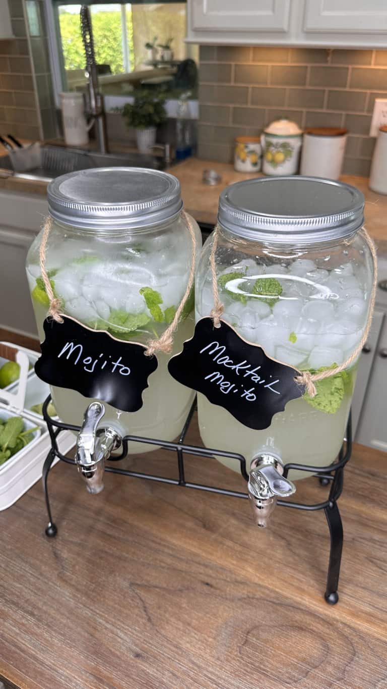 Two large glass beverage dispensers filled with ice, mint leaves, and a light drink labeled “Mojito” and “Mocktail Mojito” on black signs, sitting on a stand on a kitchen counter.