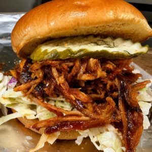 A pulled pork sandwich with barbecue sauce, shredded lettuce, and pickles on a shiny, toasted bun, served on a metal tray.
