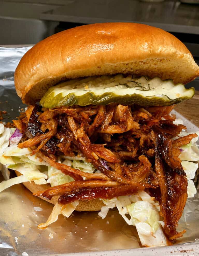 A close-up of a pulled pork sandwich with barbecue sauce, sliced pickles, and shredded lettuce on a shiny, golden bun, served on a metal tray.