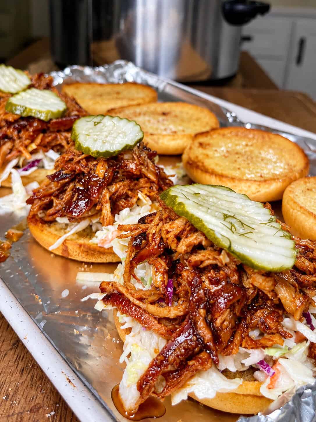 Open-faced pulled pork sandwiches topped with coleslaw and sliced pickles are arranged on a baking sheet. Toasted buns and a slow cooker are visible in the background on a kitchen counter.