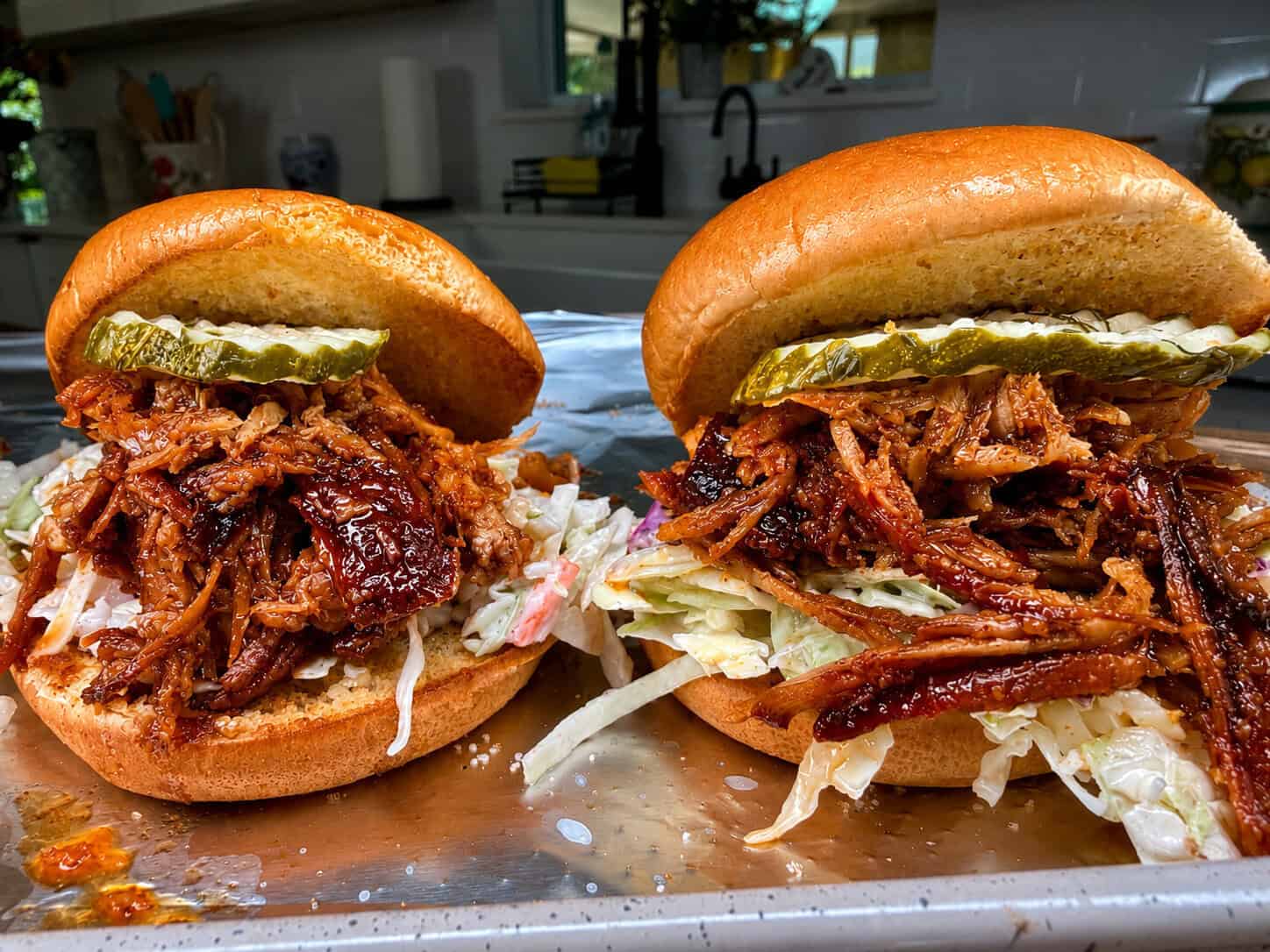 Two pulled pork sandwiches on buns with shredded lettuce, coleslaw, and pickle slices, set on a metal tray in a modern kitchen. The pork is saucy and generous, with a toasted bun top slightly tilted.