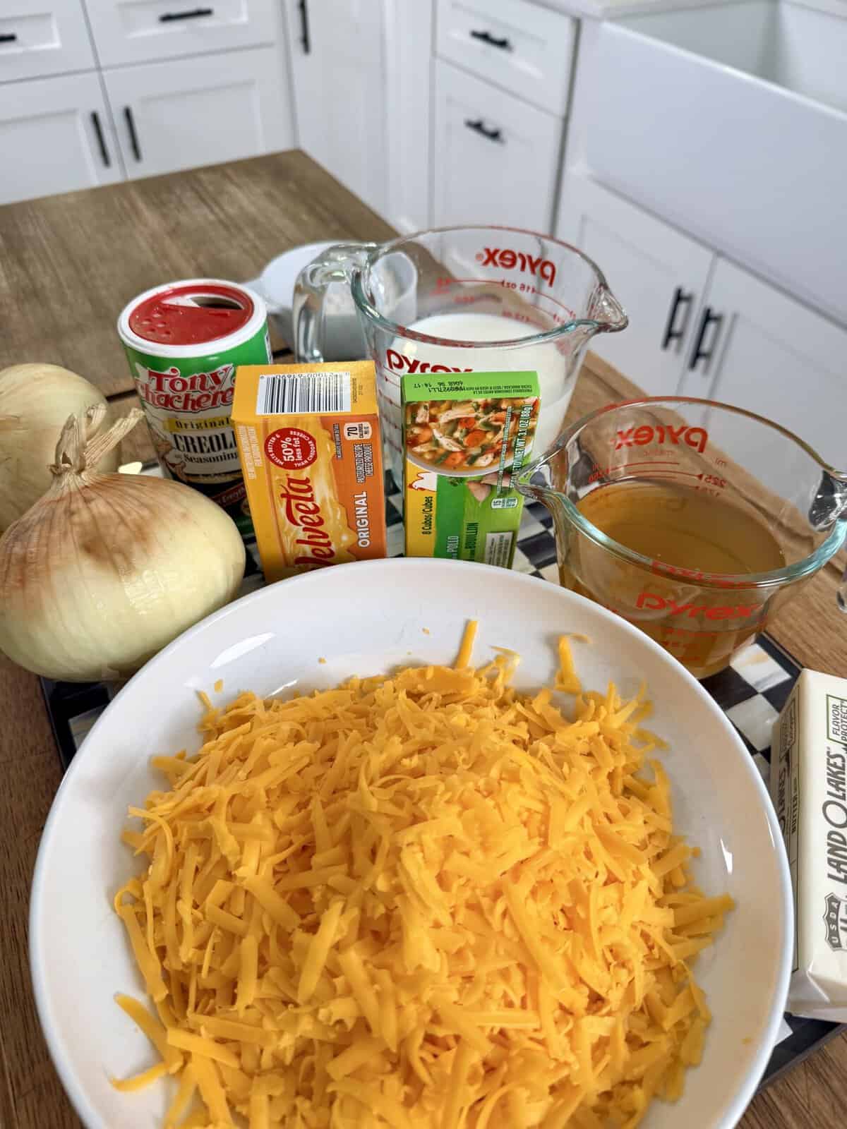 Shredded cheddar cheese, onions, butter, chicken broth, milk, sharp cheddar, a spice canister, and soup mix are arranged on a kitchen counter with white cabinets in the background.