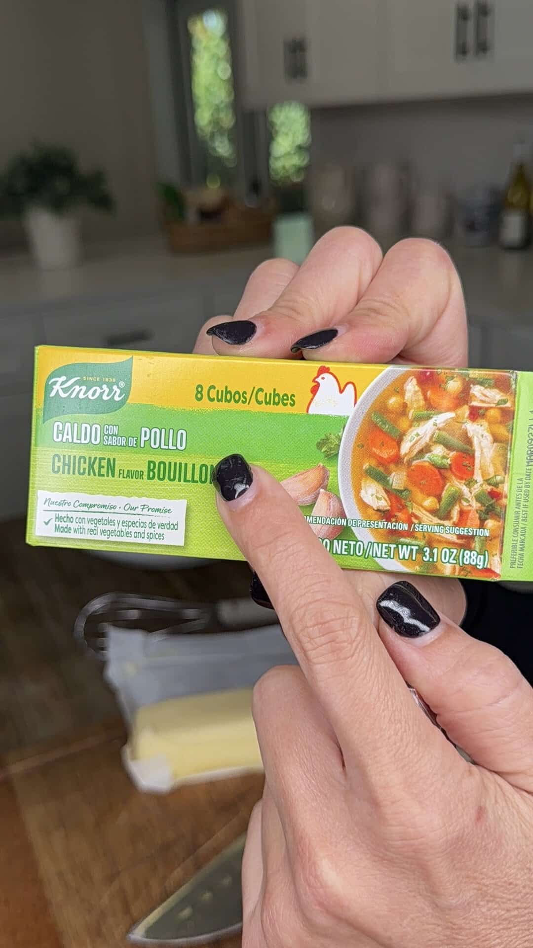 A person with black nail polish holds and points to a box of Knorr chicken flavor bouillon cubes in a kitchen.