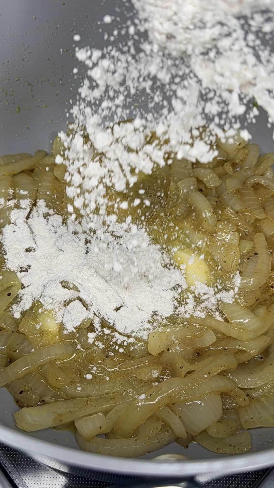 Sliced onions sautéing in a pan with melted butter, while white flour is being sprinkled over them, likely as part of a cooking or roux-making process.