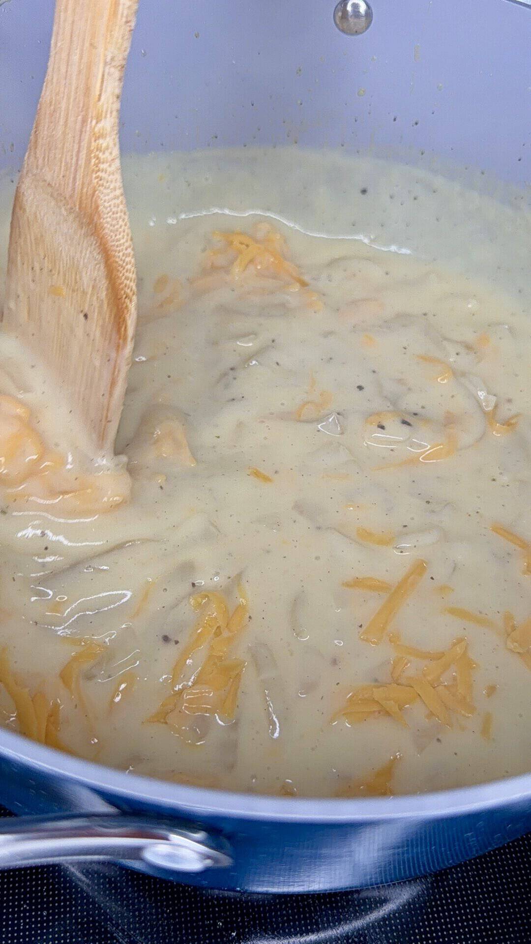 A wooden spoon stirs a creamy cheesy sauce with shredded cheese melting in a silver pot on a stovetop.