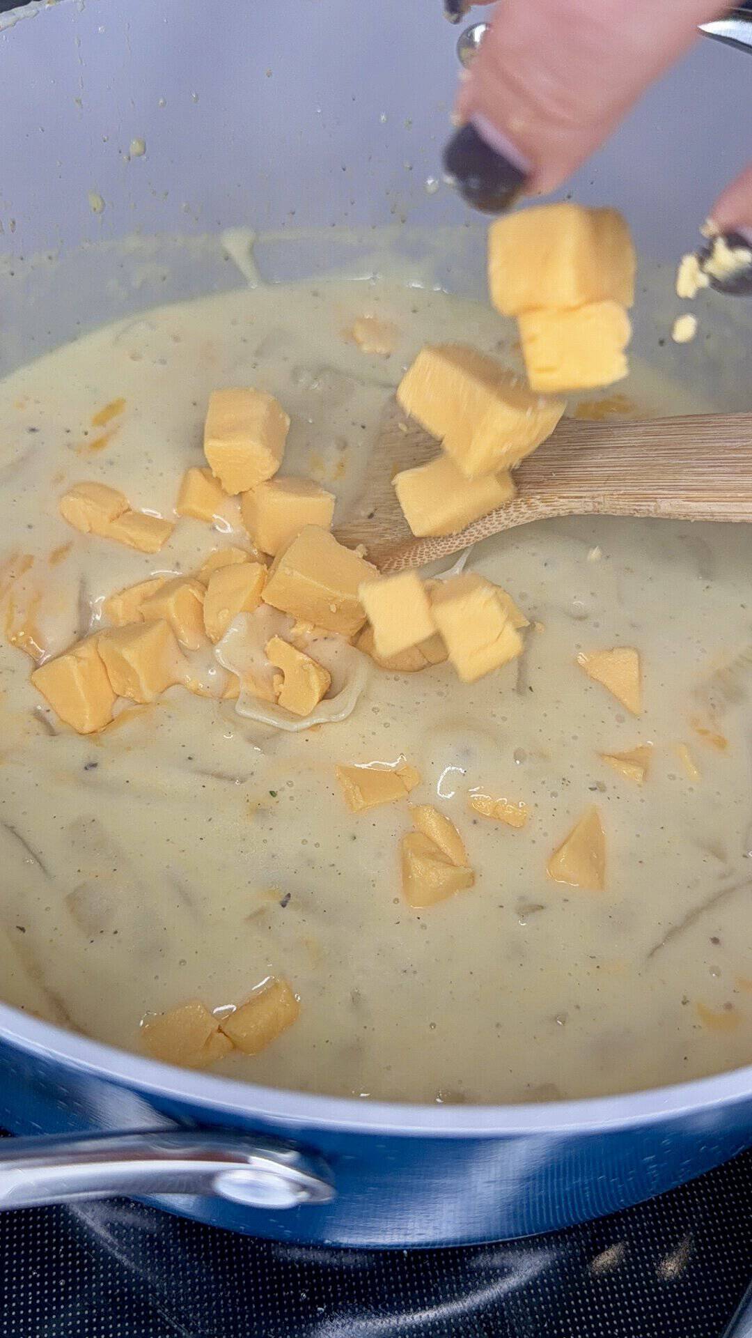 A hand drops cubes of cheddar cheese into a pot of creamy sauce while stirring with a wooden spoon.