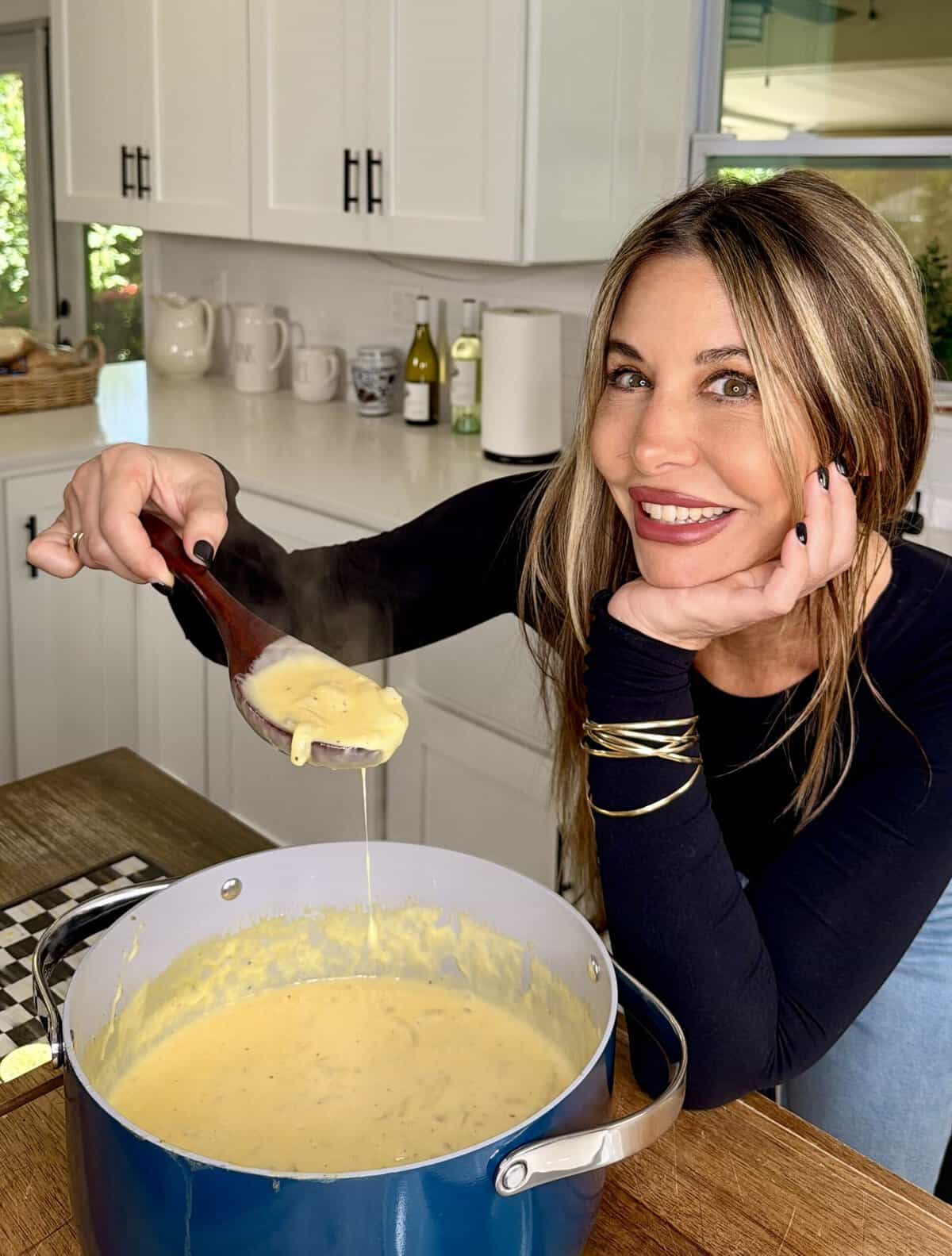 Woman in a kitchen smiling while holding a wooden spoon with creamy soup over a blue pot on the stove. White cabinets and kitchen items are visible in the background.