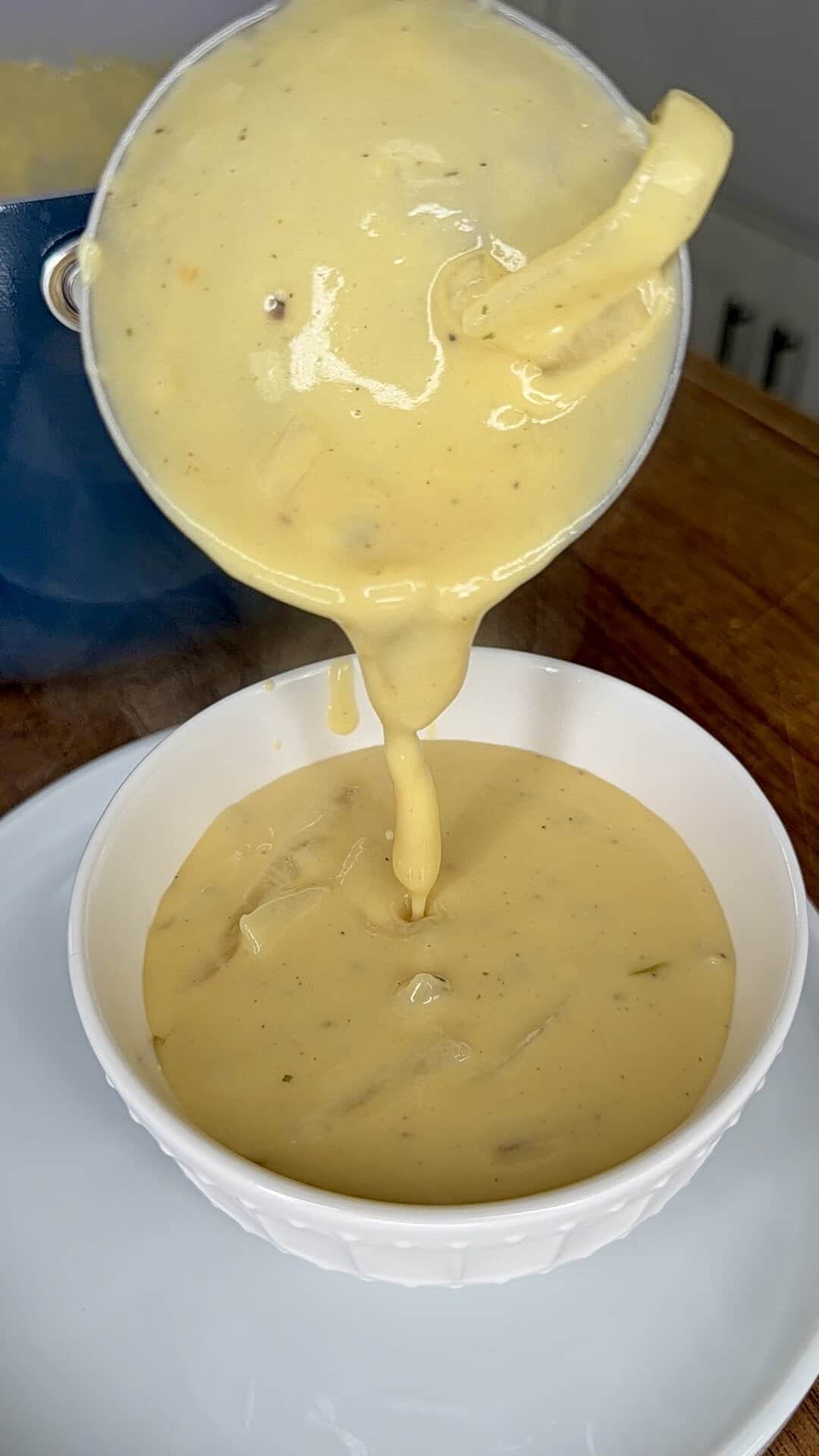 A ladle pours thick, creamy yellow sauce with visible herbs into a white bowl on a white plate, set on a wooden surface.