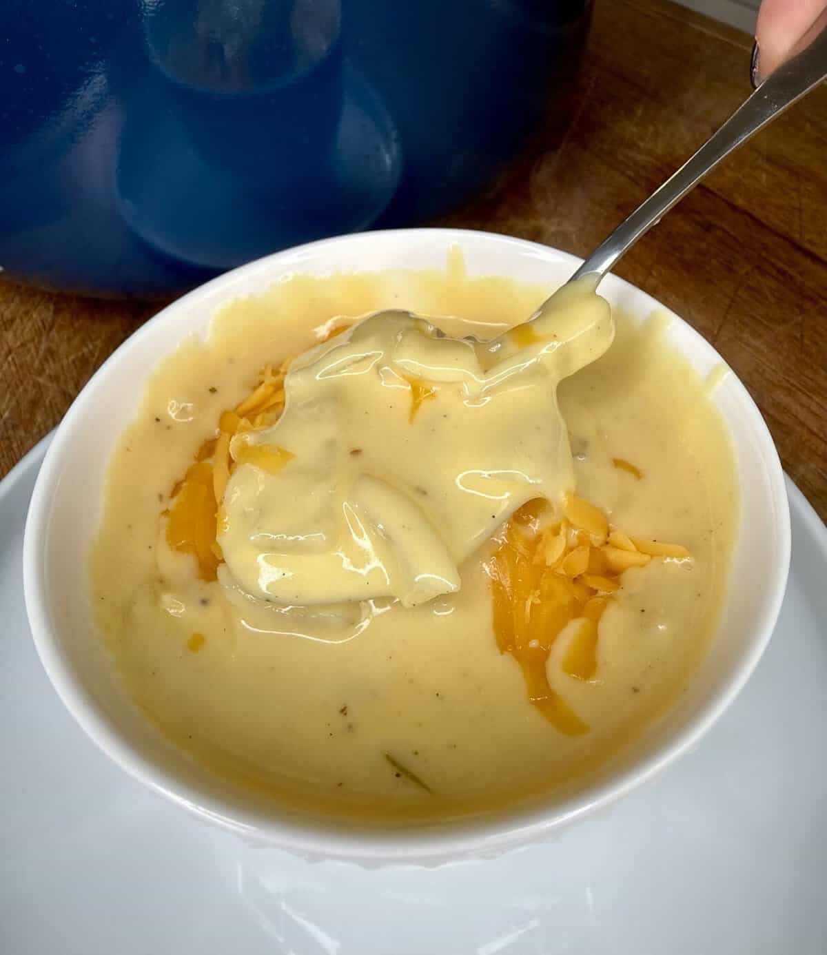 A bowl of creamy yellow soup topped with shredded cheddar cheese, with a spoon lifting a portion. A blue pot is visible in the background on a wooden surface.