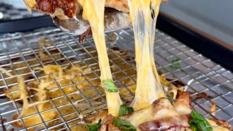 A close-up of cheesy baked chicken being lifted from a wire rack, showing melted cheddar cheese stretching, topped with chopped green onions. The background features a kitchen setting.