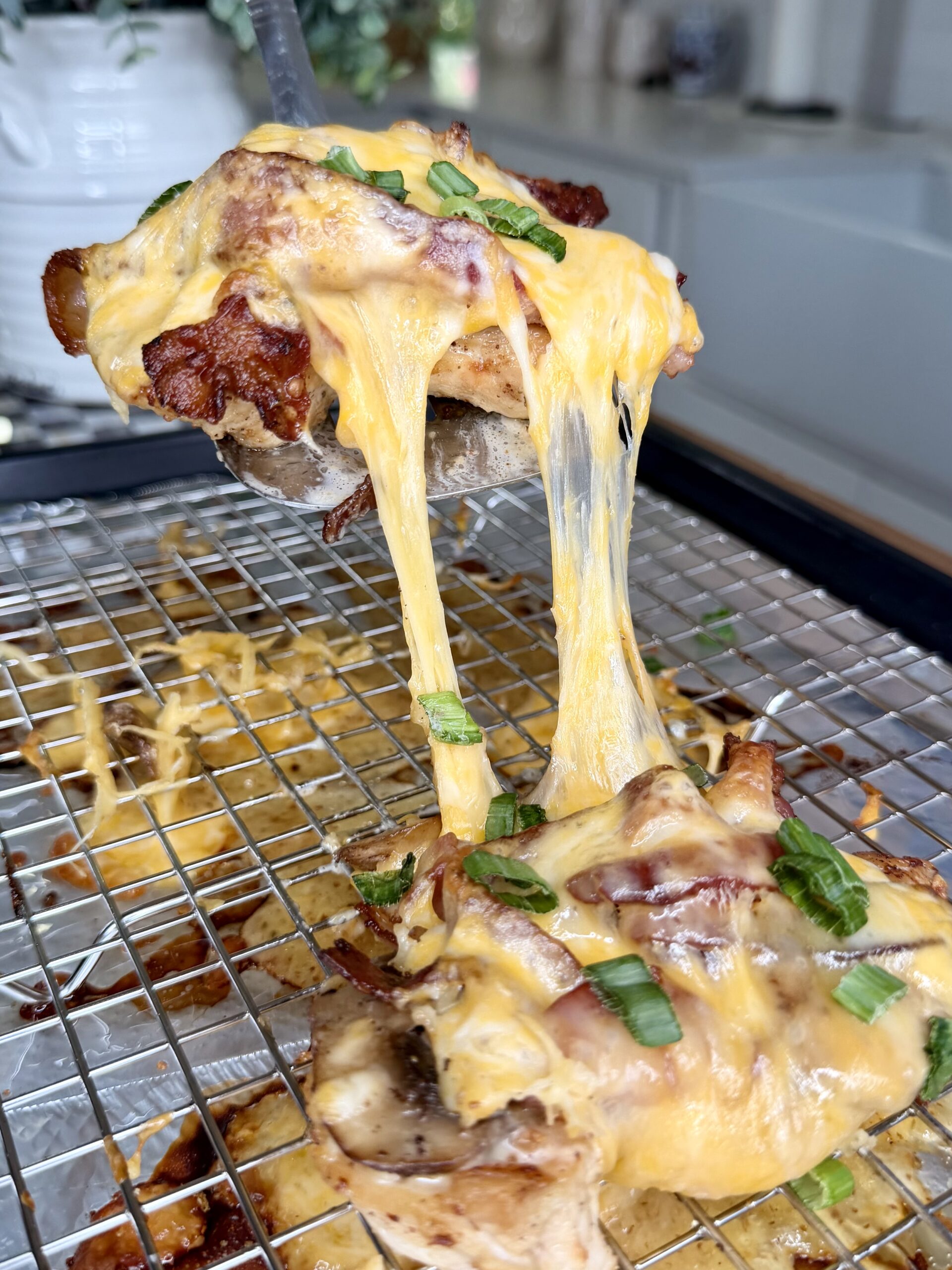 A close-up of cheesy baked chicken being lifted from a wire rack, showing melted cheddar cheese stretching, topped with chopped green onions. The background features a kitchen setting.