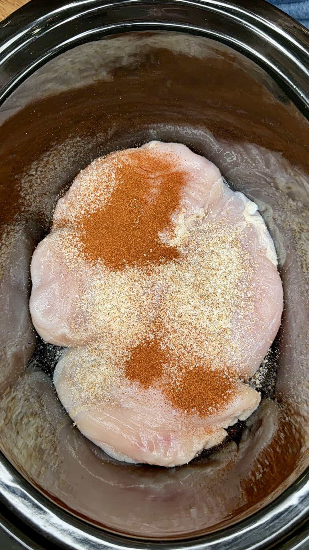 Raw chicken breasts in a slow cooker, topped with paprika and what appears to be garlic or onion powder, ready to be cooked.
