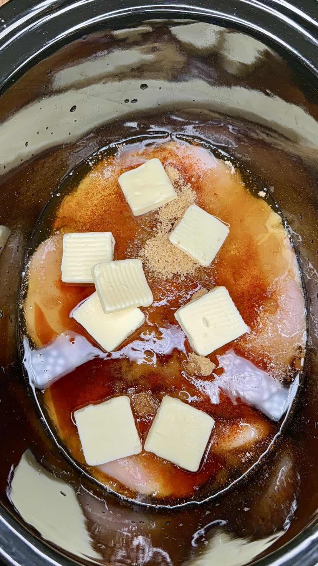 Raw chicken breasts in a slow cooker, topped with slices of butter, brown sugar, and a dark sauce, ready for cooking.