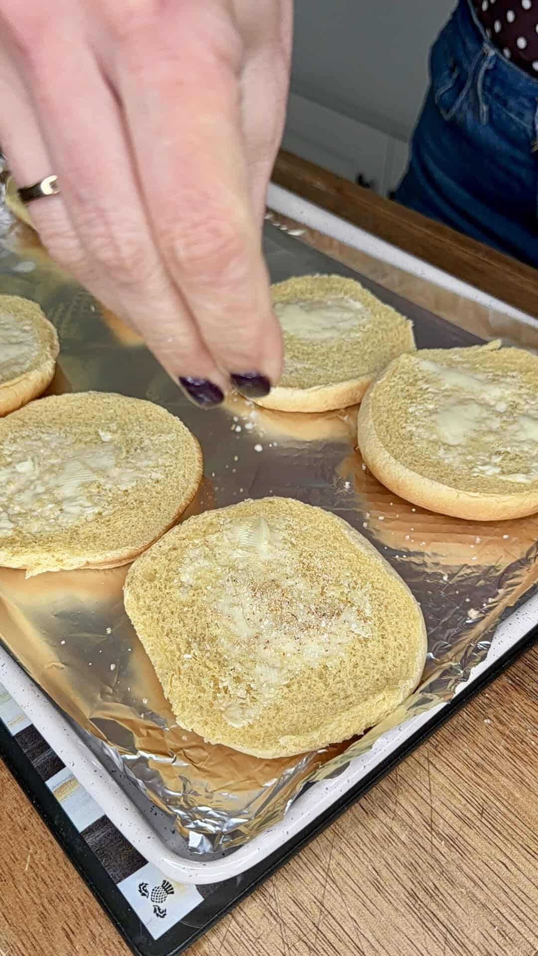 A hand sprinkles seasoning onto buttered hamburger buns arranged on a foil-lined baking sheet, ready for toasting.