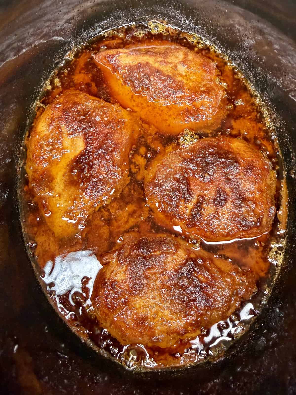 Four pieces of seasoned, cooked chicken breast sit in a slow cooker, surrounded by a reddish-brown sauce and spices. The chicken appears tender with a slightly crispy, flavorful coating.