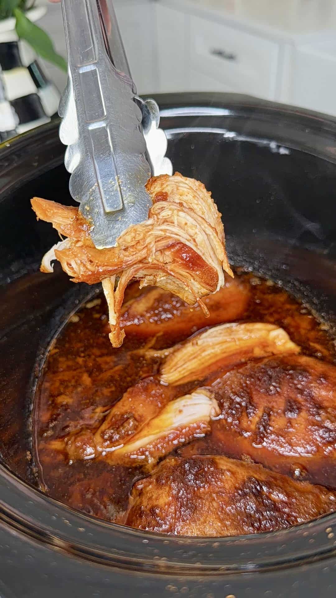 Metal tongs hold up shredded, saucy chicken over a slow cooker filled with more cooked, seasoned chicken in rich, dark sauce. Kitchen countertop and cabinets are visible in the background.