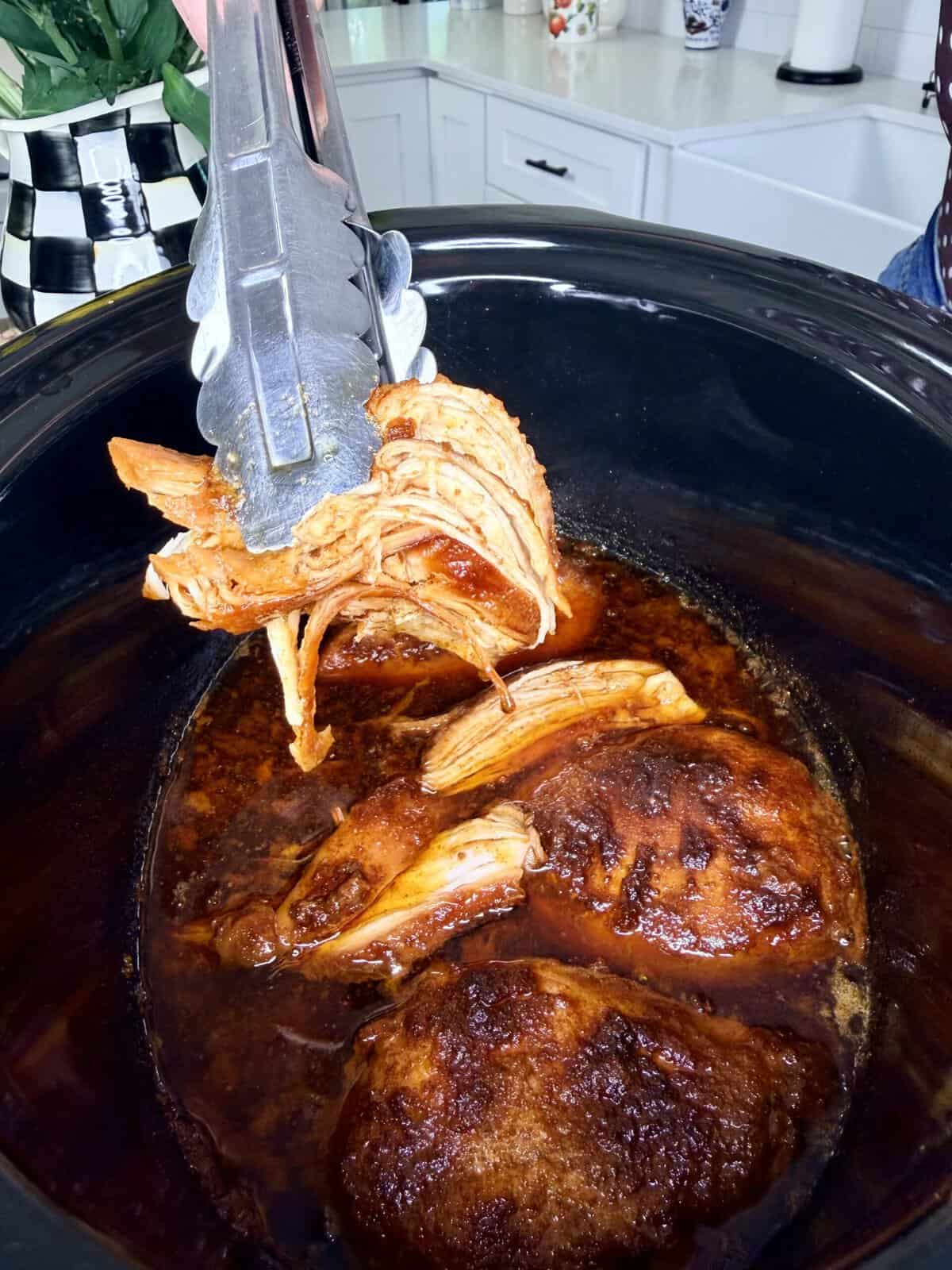 Tongs lifting shredded, cooked BBQ chicken from a slow cooker filled with juicy, saucy chicken pieces in a kitchen setting.