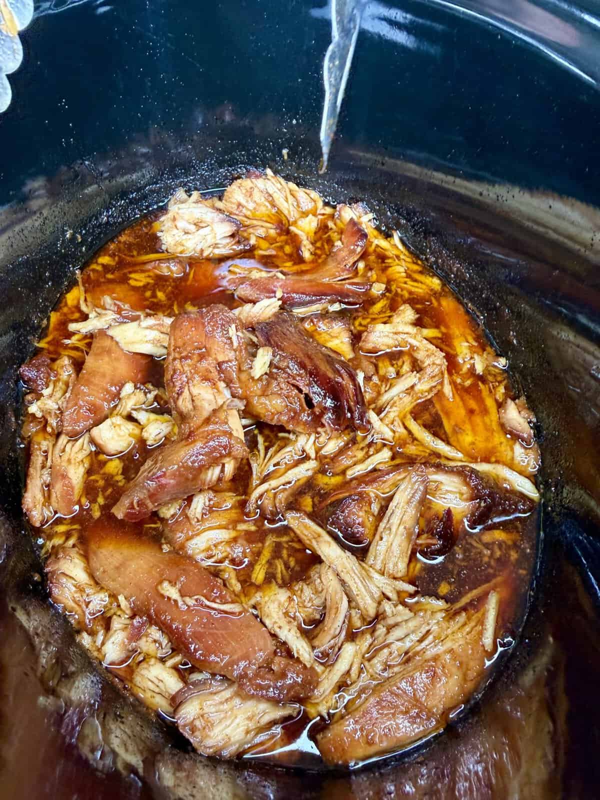 Shredded pork in a dark, flavorful sauce inside a slow cooker, with pieces of tender meat soaking in the juices.
