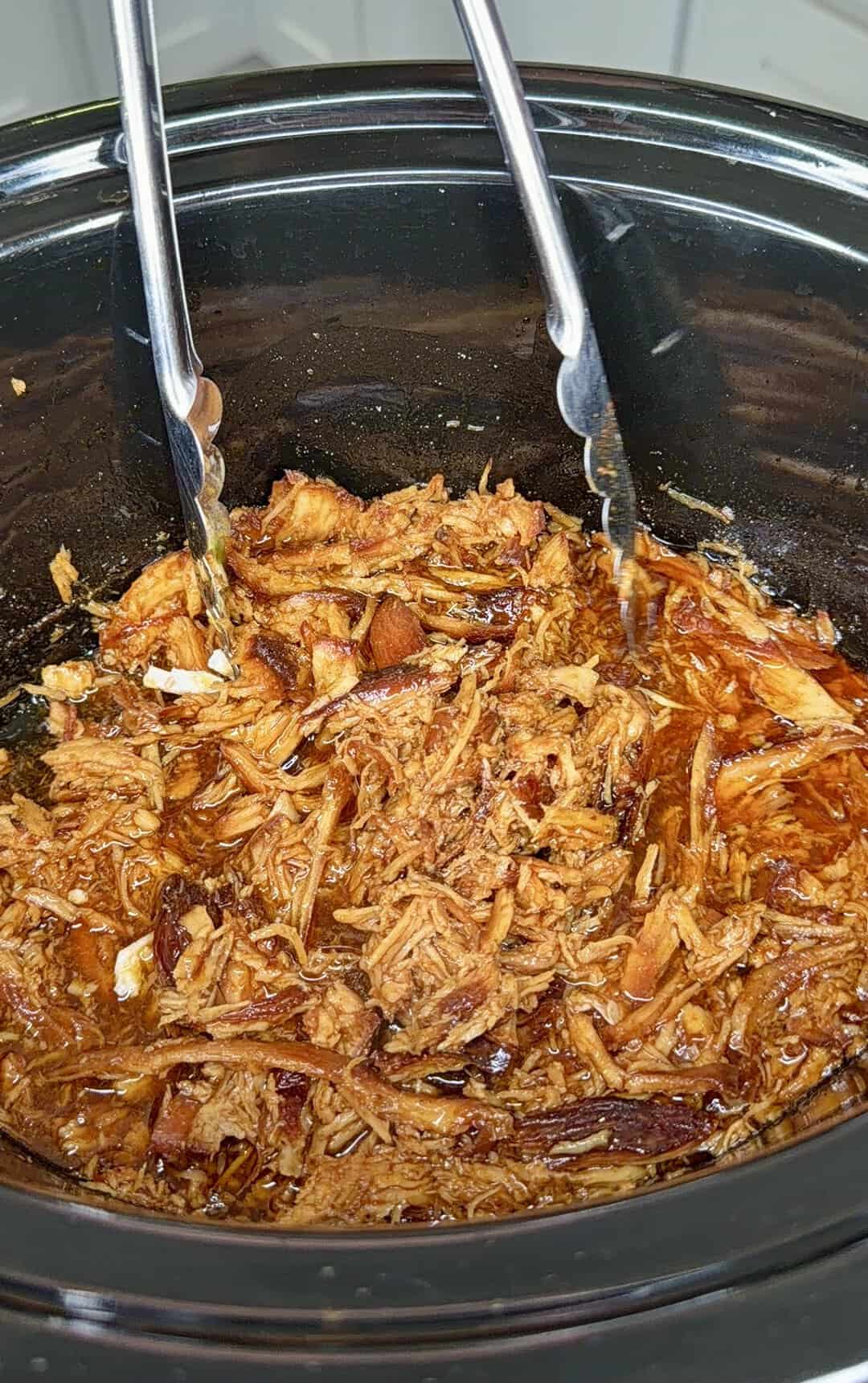 Shredded barbecue chicken in a slow cooker with metal tongs resting inside, the chicken is mixed with a rich, reddish sauce.